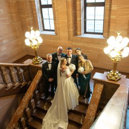 NIKA KHRYSTYNA NEDOMOLKINA - Panna młoda w białej sukni i panowie w smokingach na marmurowych schodach z kwiatami, widok z góry, oświetlenie lampami w stylu retro, duże okna w tle.