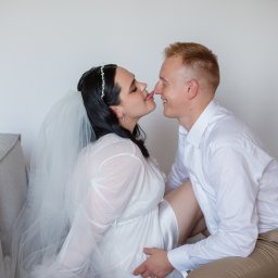 NIKA KHRYSTYNA NEDOMOLKINA - Uśmiechnięta para w jasnych strojach siedzi blisko, dotykając się nosami. Kobieta w welonie i sukience, mężczyzna w koszuli. Delikatne, naturalne światło.