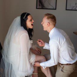 NIKA KHRYSTYNA NEDOMOLKINA - Radosna panna młoda w bieli siedzi, a pan młody klęczy, zakładając jej podwiązkę. Uśmiechnięci, w jasnym wnętrzu z minimalistycznymi obrazami w tle.