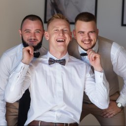 NIKA KHRYSTYNA NEDOMOLKINA - Trzech uśmiechniętych mężczyzn w białych koszulach i muchach, pozujących do zdjęcia w jasnym wnętrzu. Środkowy mężczyzna ma wzorzystą muchę.