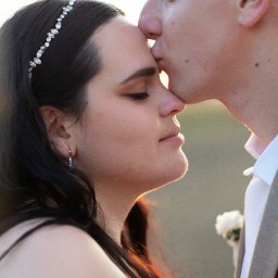 NIKA KHRYSTYNA NEDOMOLKINA - Profil narzeczonej z diademem, całowanej w czoło przez narzeczonego w beżowym garniturze, w ciepłym świetle zachodzącego słońca. Delikatne kolczyki i kwiat w butonierce.