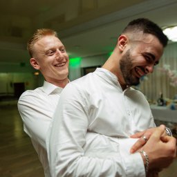 NIKA KHRYSTYNA NEDOMOLKINA - Radosne ujęcie dwóch mężczyzn w białych koszulach, śmiejących się podczas uroczystości. W tle widoczny stół z dekoracjami i kobieta w zielonej sukience.