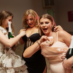 NIKA KHRYSTYNA NEDOMOLKINA - Trzy kobiety w sukienkach świętują z szampanem i pieniędzmi na imprezie. Jedna z nich trzyma butelkę, a pozostałe mają banknoty w dekoltach. W tle widoczny sprzęt nagłośnieniowy.