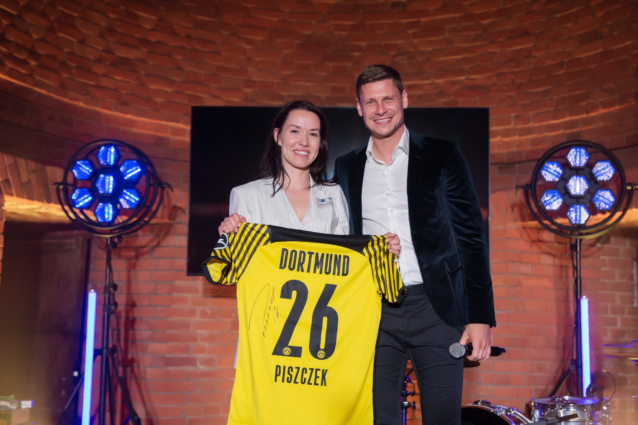 Uśmiechnięta kobieta i mężczyzna prezentują żółtą koszulkę piłkarską z autografem na tle ceglanej ściany z oświetleniem eventowym.