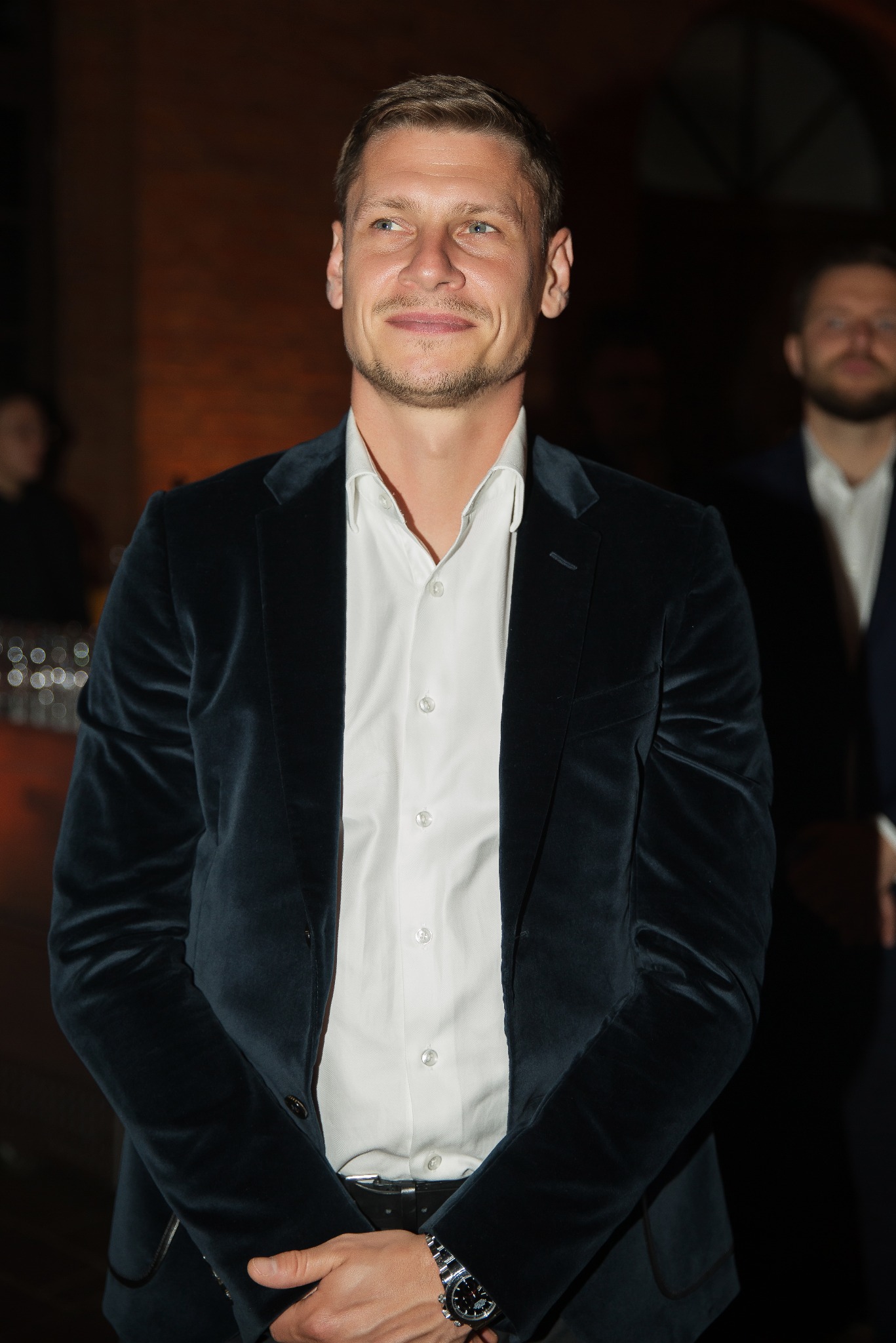 Mężczyzna w aksamitnej marynarce i białej koszuli, z zegarkiem na nadgarstku, uśmiecha się w kierunku obiektywu. W tle widoczna inna osoba i elementy dekoracyjne.