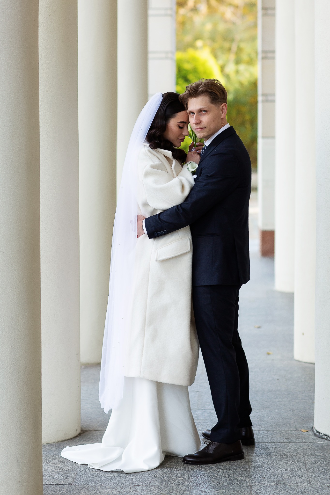 Elegancka para młoda w objęciach, on w ciemnym garniturze, ona w białym płaszczu i welonie, na tle kolumn. Subtelne światło podkreśla uroczysty charakter.
