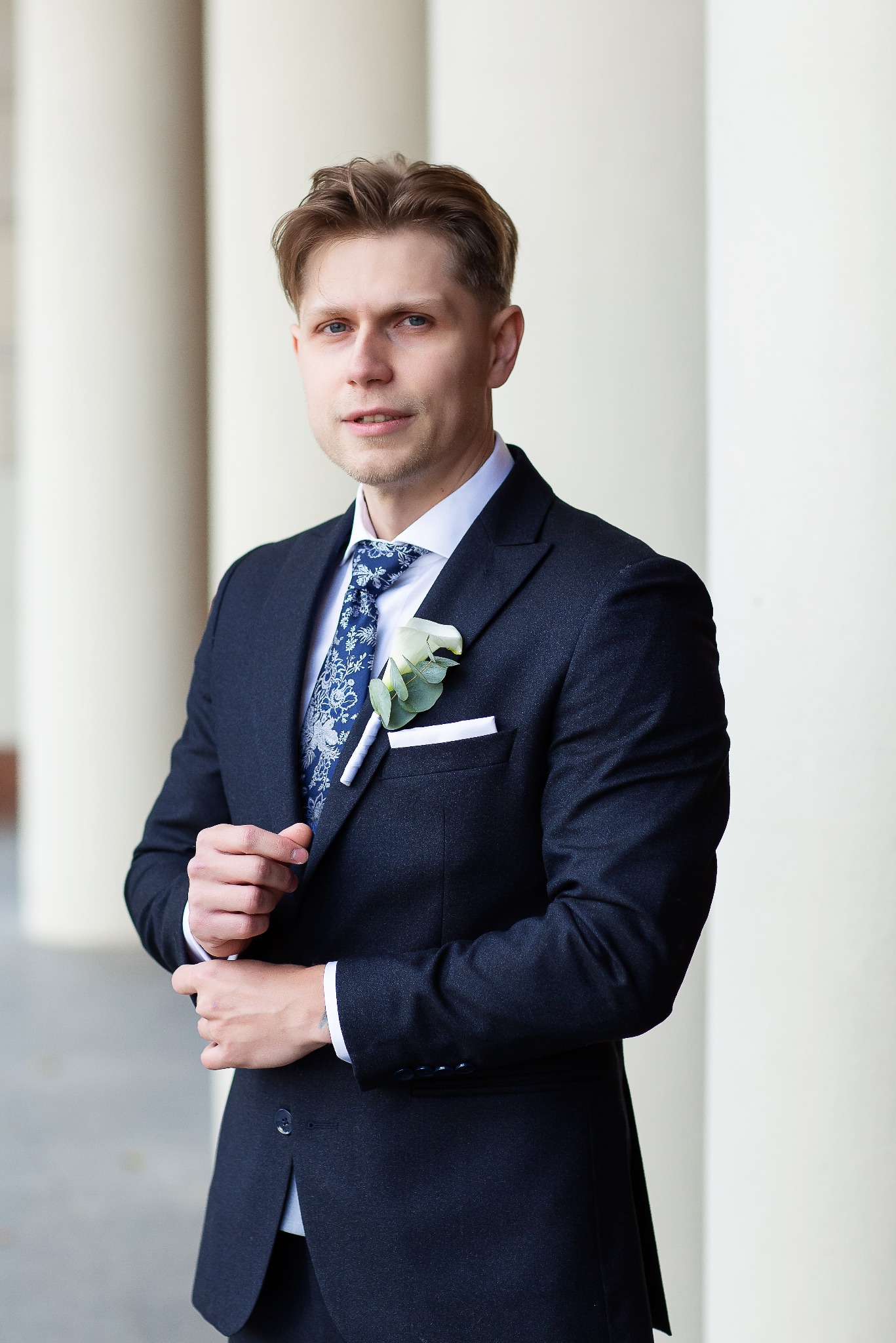 Elegancki portret mężczyzny w granatowym garniturze z butonierką i wzorzystym krawatem, pozującego na tle kolumn. Delikatny uśmiech, dopasowany strój.
