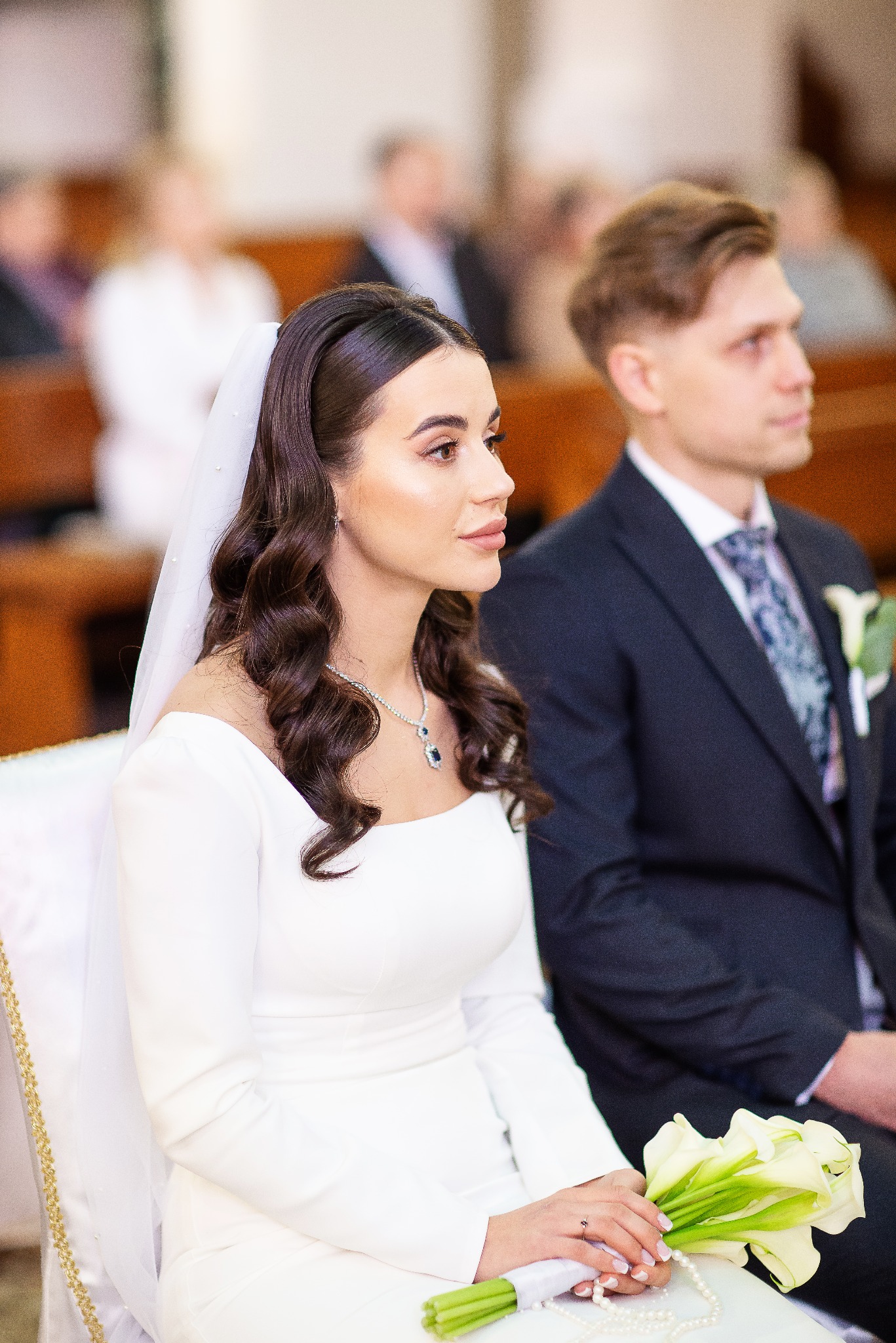 Elegancka panna młoda w białej sukni i welonie, siedząca obok pana młodego w kościele, trzyma bukiet kalii. W tle widoczni goście.