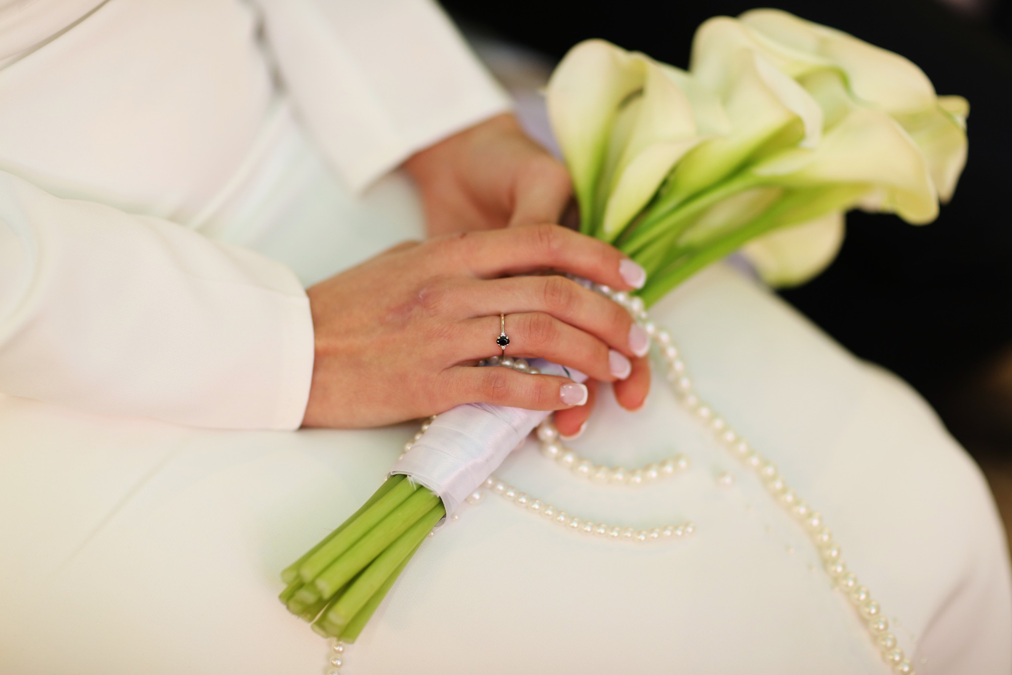 Dłonie panny młodej z pierścionkiem zaręczynowym trzymają bukiet białych kalii, ozdobiony perłami. Ujęcie z bliska, skupione na detalu.