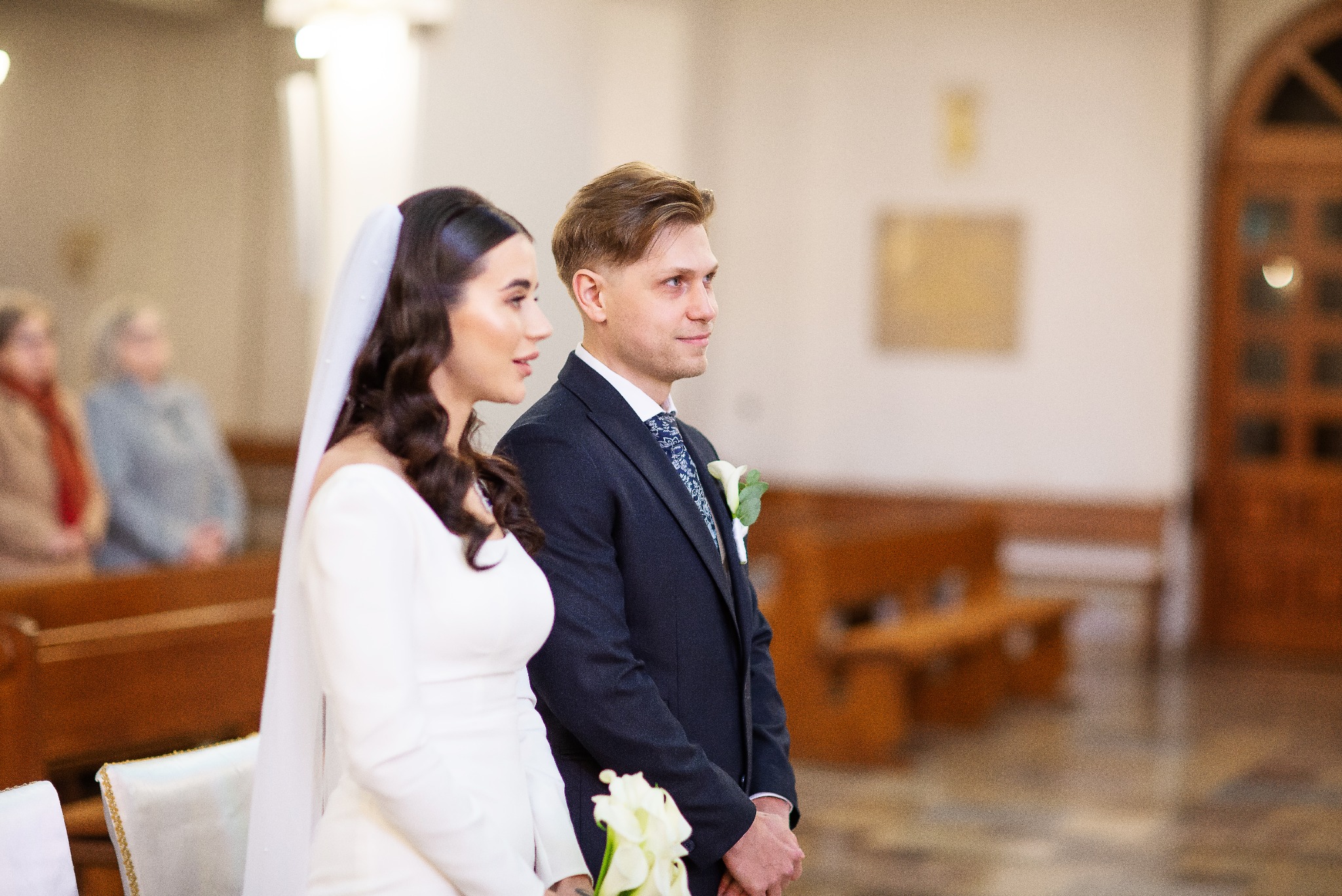 Para młoda w kościele podczas ceremonii ślubnej. Panna młoda w białej sukni z welonem, pan młody w granatowym garniturze. W tle ławki i drewniane drzwi.