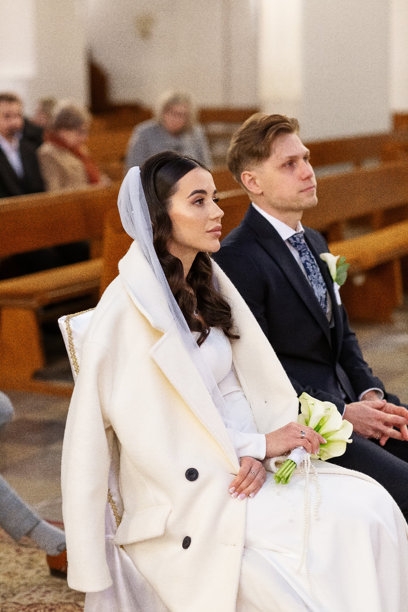 Elegancka para młoda siedzi w kościele podczas ceremonii ślubnej. Panna młoda w białym płaszczu i welonie, pan młody w ciemnym garniturze. W tle ławki i inni goście.