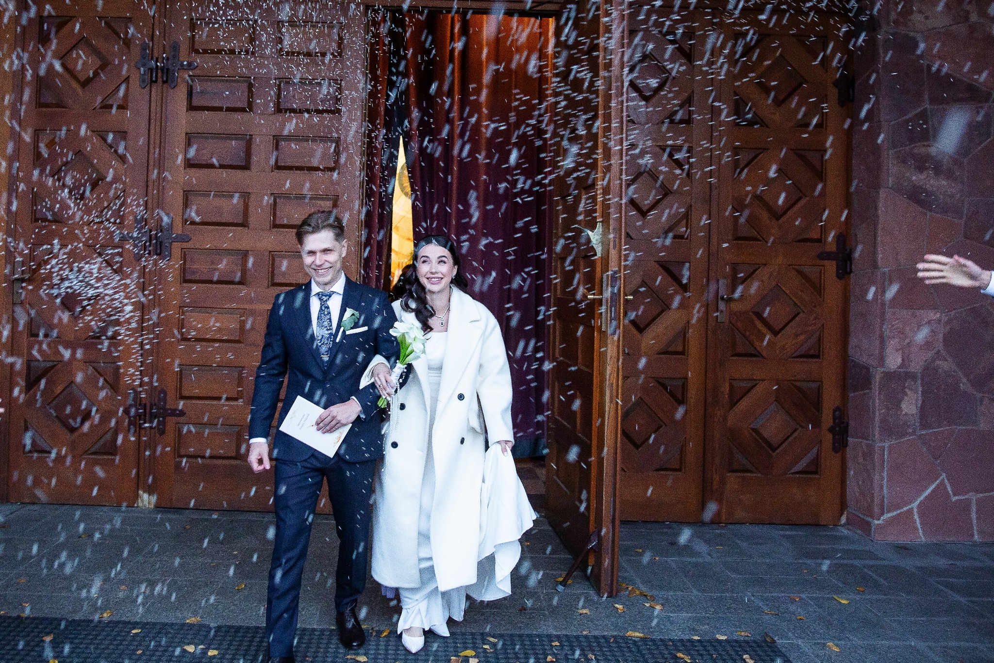 Uśmiechnięta para młoda wychodzi z kościoła, obsypywana konfetti na tle masywnych, drewnianych drzwi. Panna młoda w białym płaszczu, pan młody w granatowym garniturze.