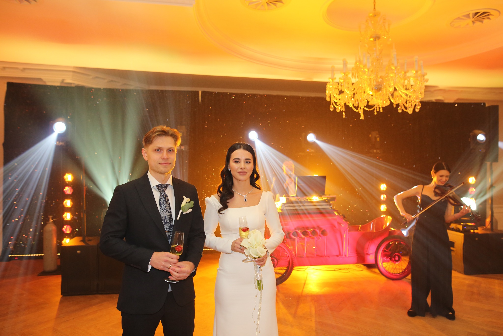 Elegancka para młoda z kieliszkami szampana na tle sceny z oświetlonym, różowym samochodem i grającą na skrzypcach kobietą. Dekoracja sali z żyrandolem.