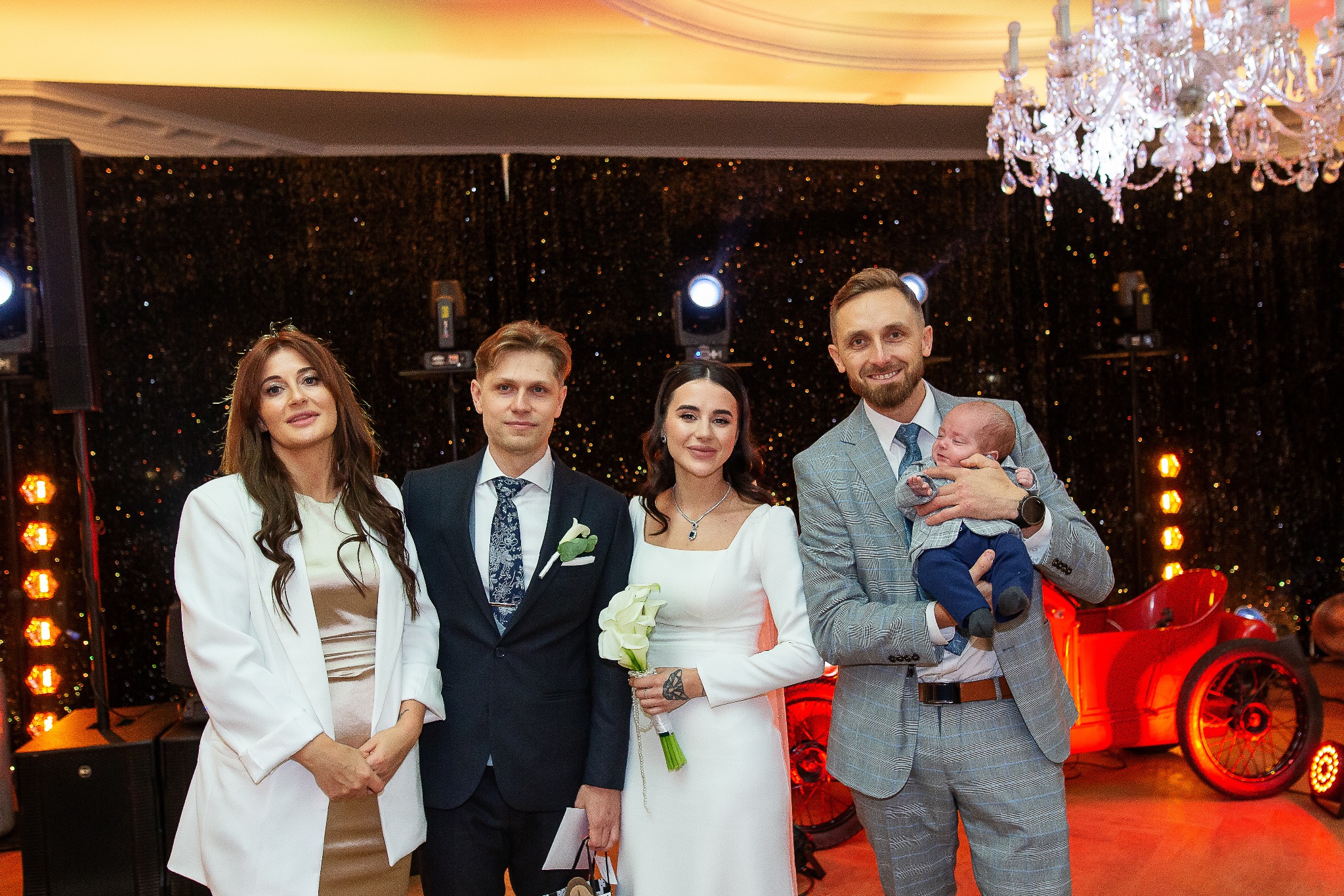 Elegancka sesja rodzinna w studio: rodzice z noworodkiem i goście pozują na tle dekoracji z czerwonym pojazdem i błyszczącej ściany. W tle kryształowy żyrandol.