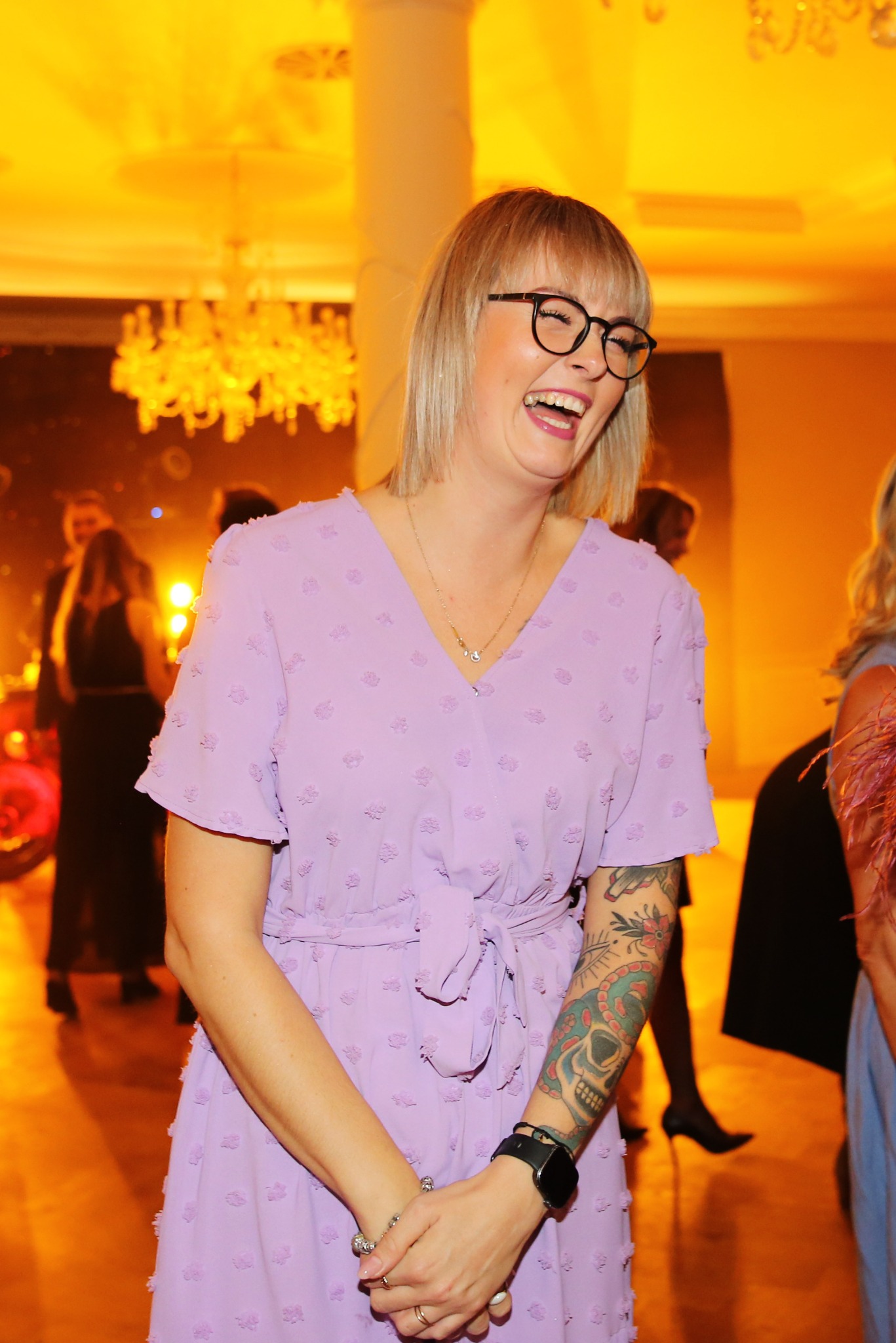 Uśmiechnięta blondynka w okularach i liliowej sukience na tle sali balowej z żyrandolem. Widoczny tatuaż na ramieniu i zegarek. Ciepłe oświetlenie.