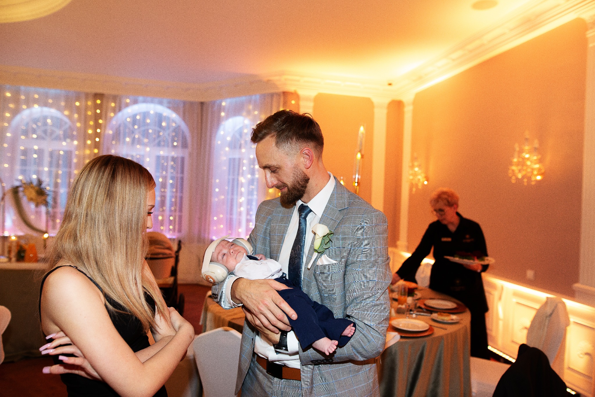 Elegancki mężczyzna w garniturze trzyma śpiące niemowlę w słuchawkach na tle sali bankietowej z dekoracjami świetlnymi i gośćmi. Ujęcie z uroczystości rodzinnej.
