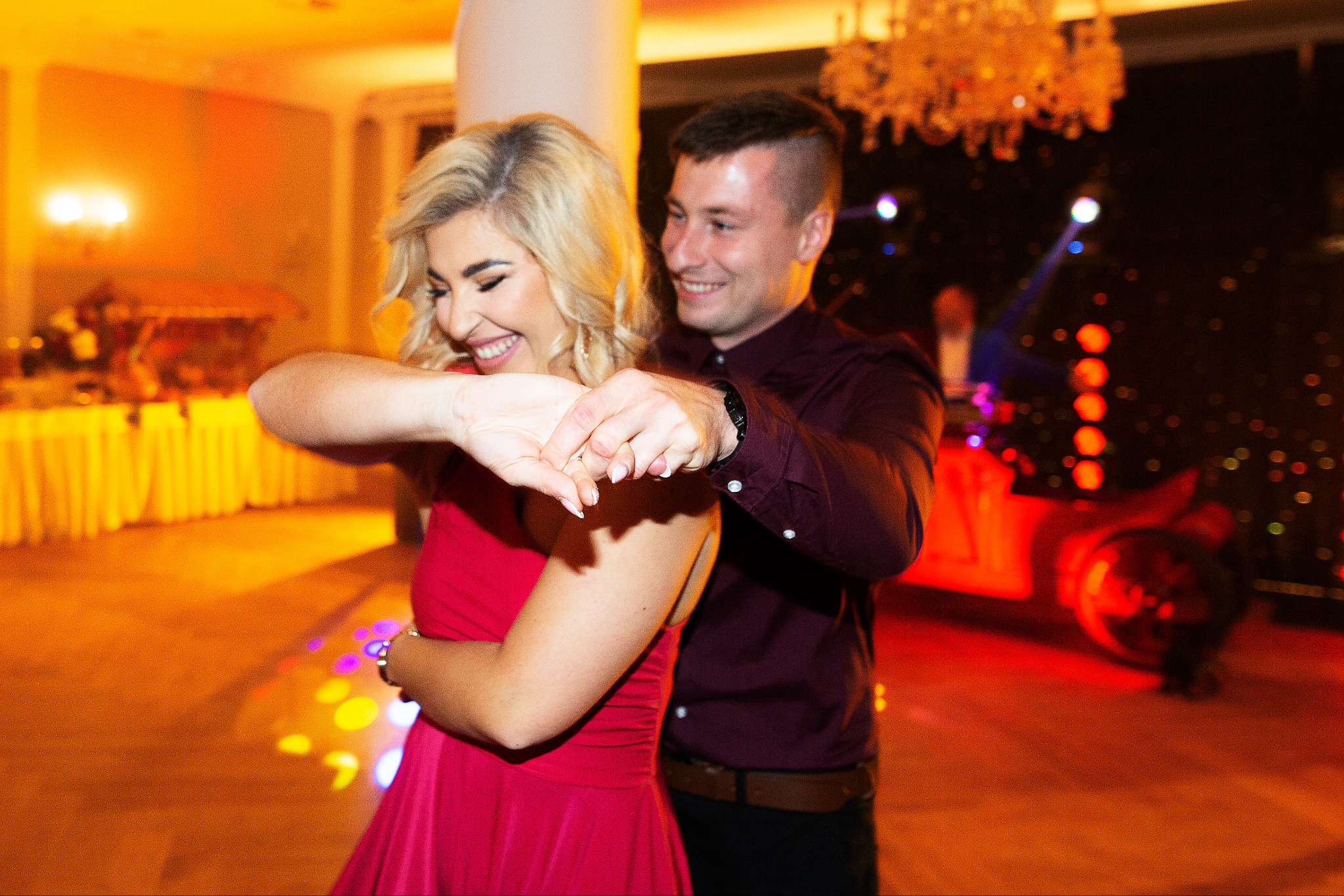Uśmiechnięta para tańczy na parkiecie podczas eventu. Kobieta w czerwonej sukience, mężczyzna w bordowej koszuli. W tle dekoracje i oświetlenie imprezowe.