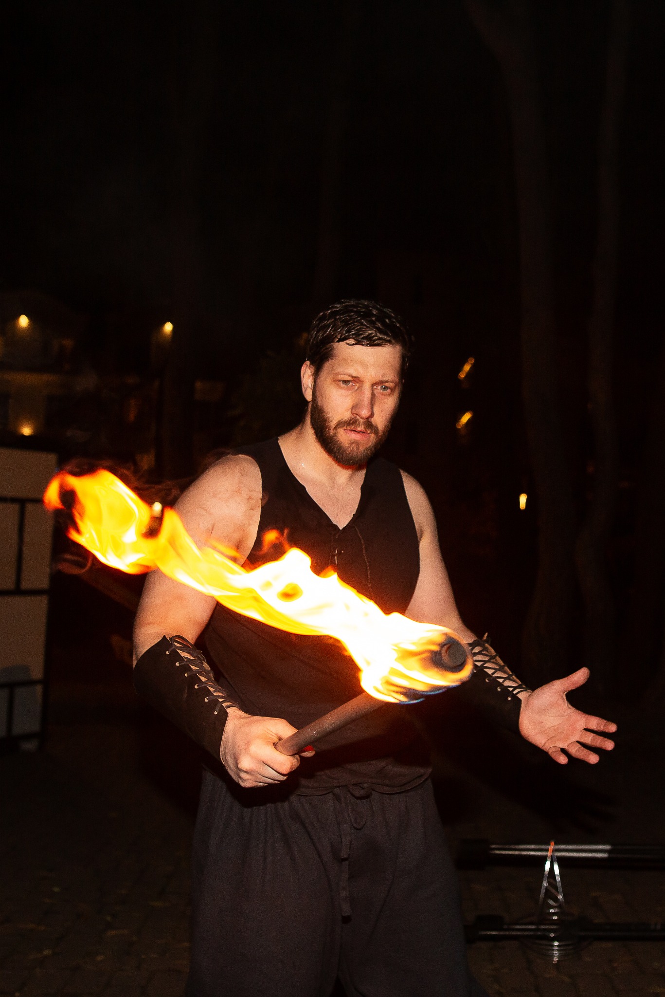 Mężczyzna z brodą w czarnej koszulce i skórzanych mankietach trzyma płonącą pochodnię na tle ciemności, prezentując pokaz ognia.