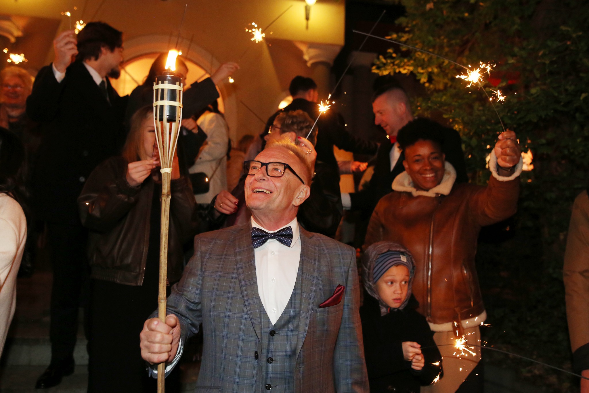 Uroczystość w Poznaniu: mężczyzna w garniturze trzyma płonącą pochodnię, w tle uśmiechnięci ludzie z zimnymi ogniami, tworząc radosną atmosferę.
