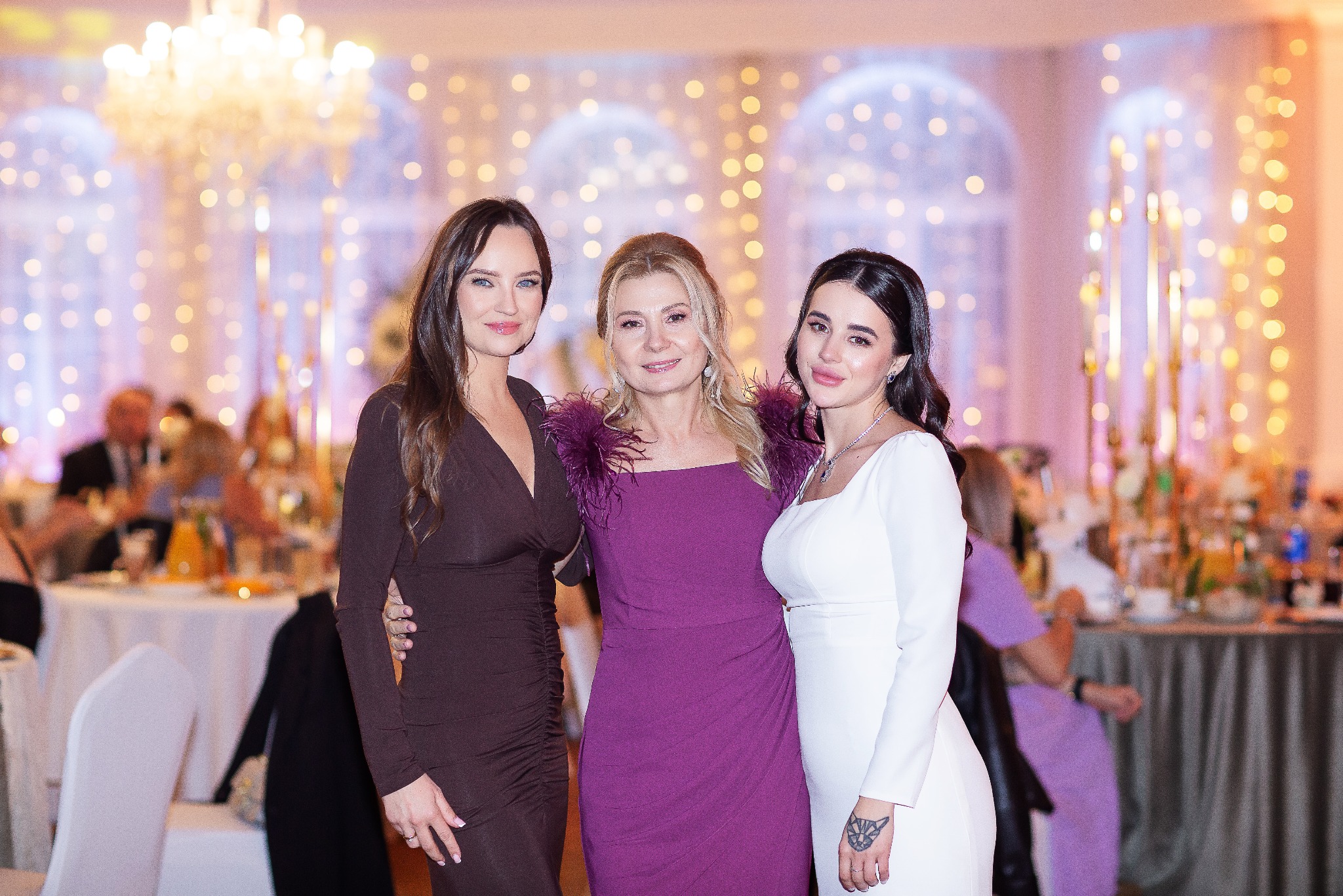 Portret trzech eleganckich kobiet w sukniach wieczorowych na tle sali balowej z dekoracjami świetlnymi i stołami. Uśmiechnięte, pozują do zdjęcia.