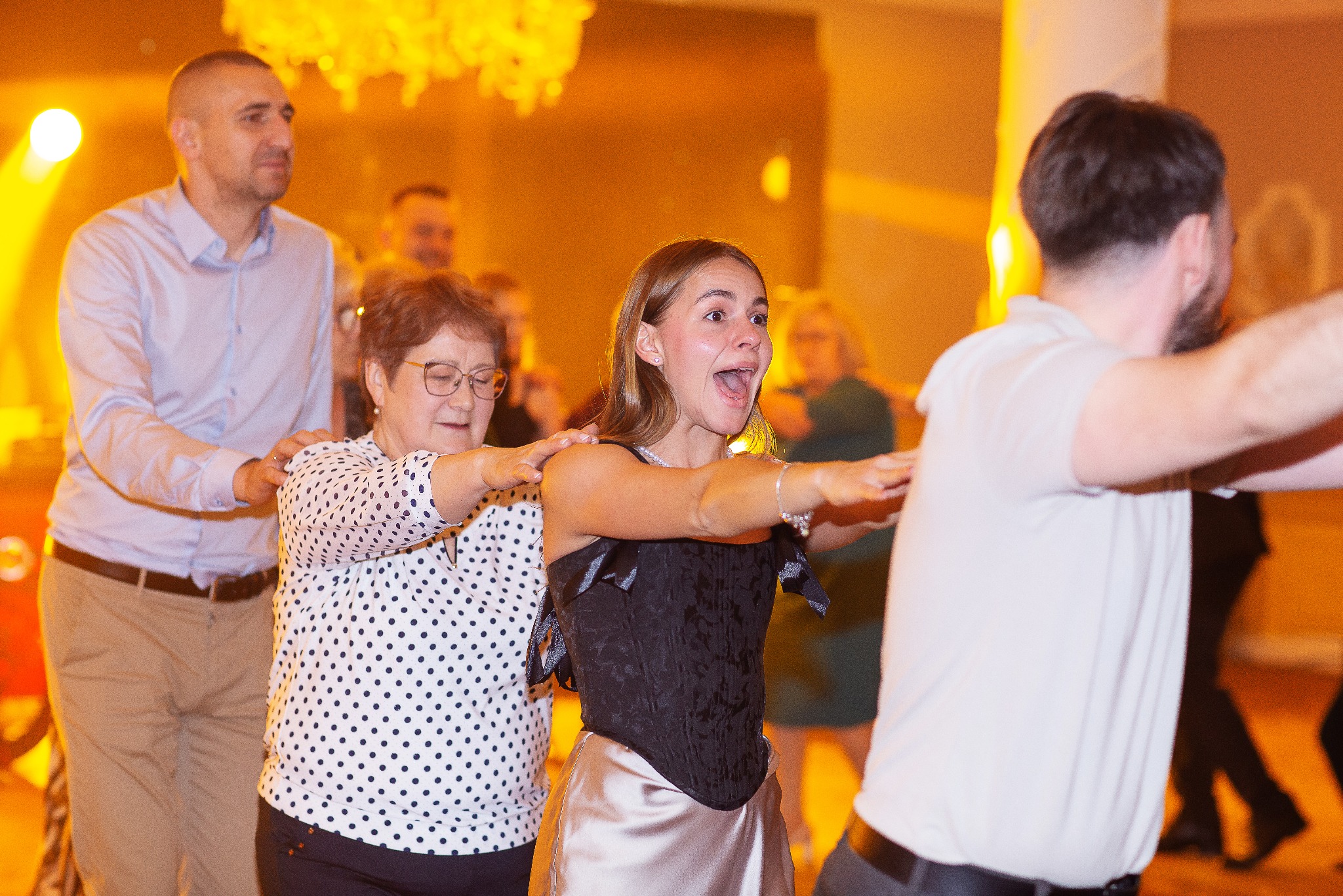 Rozbawieni goście w trakcie tańca na parkiecie, oświetlonym ciepłym światłem. Kobieta w gorsecie z szeroko otwartymi ustami, w tle mężczyzna w koszuli i kobieta w okularach.
