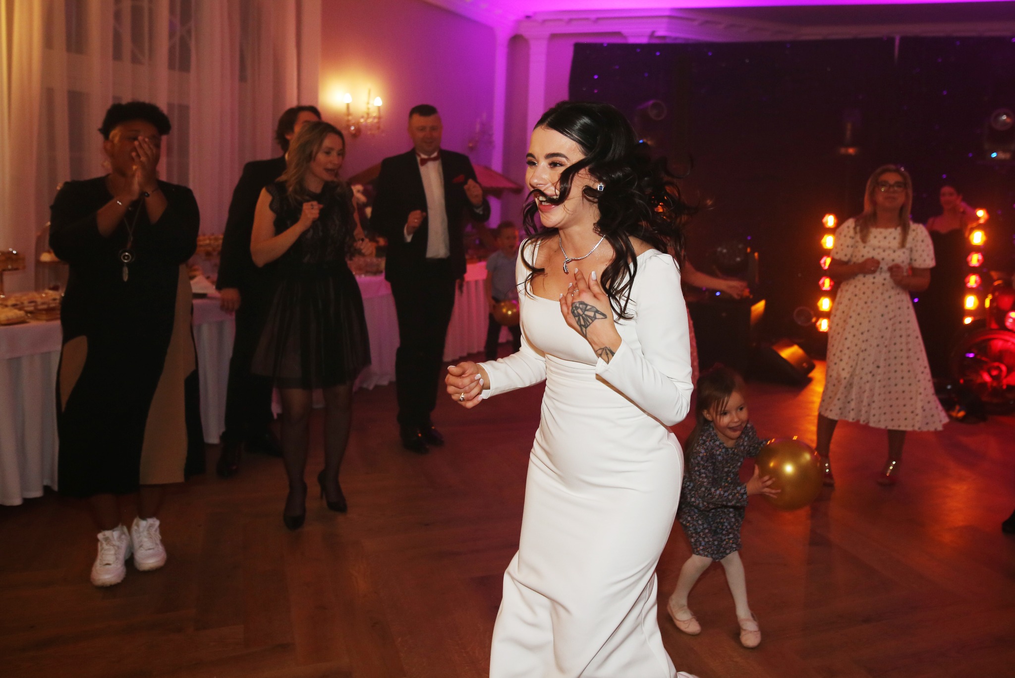 Uśmiechnięta panna młoda w białej sukni tańczy na parkiecie podczas wesela, w tle goście i dekoracje świetlne.