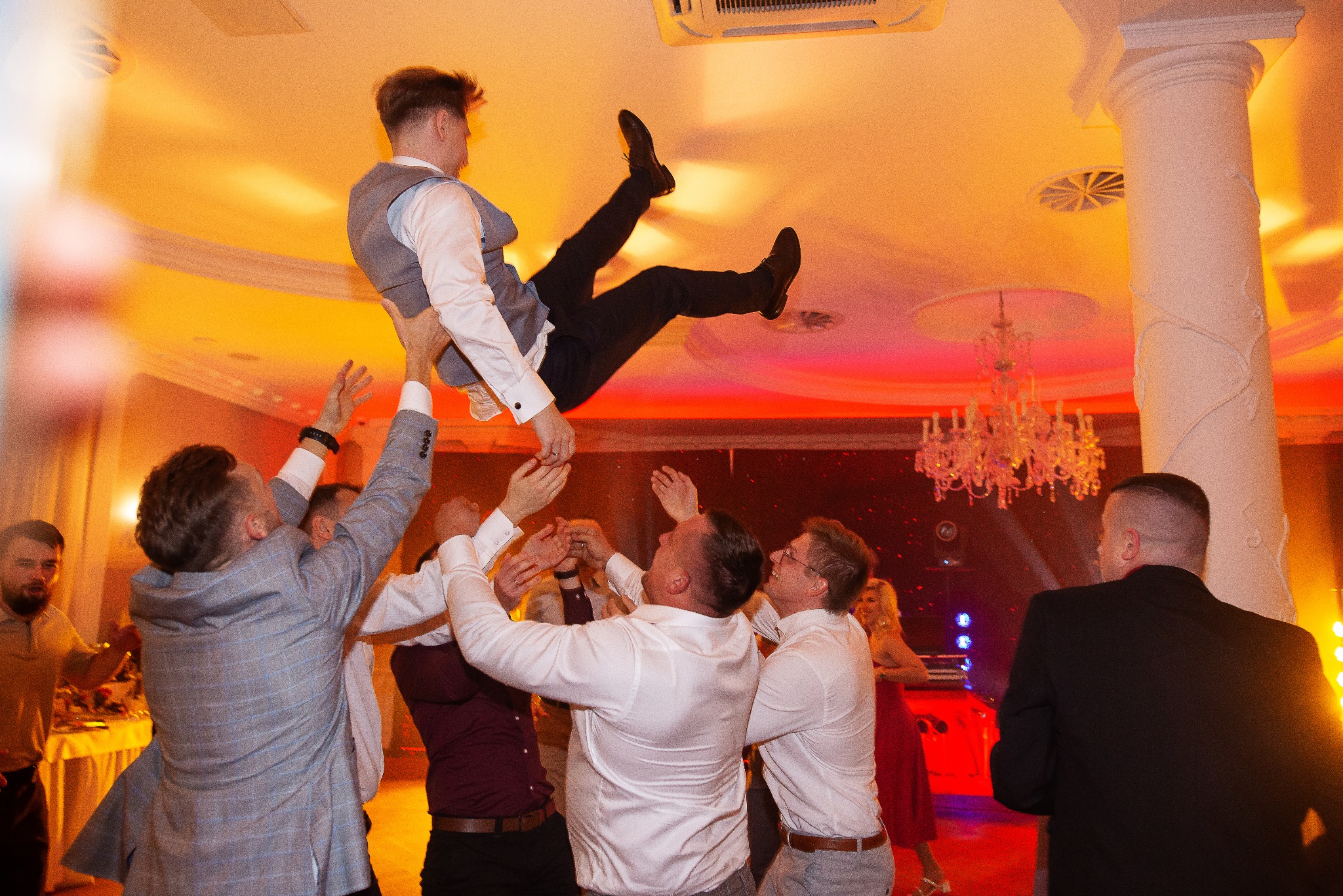 Radosna zabawa weselna: goście podrzucają pana młodego na tle kolumny i żyrandola. Dynamiczne ujęcie z parkietu, ciepłe oświetlenie podkreśla uroczysty charakter.