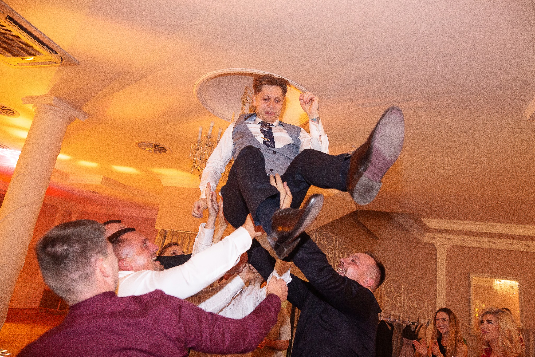 Radosny pan młody w garniturze uniesiony w górę przez gości weselnych podczas tradycyjnego tańca na parkiecie. W tle kolumny i dekoracje sali.