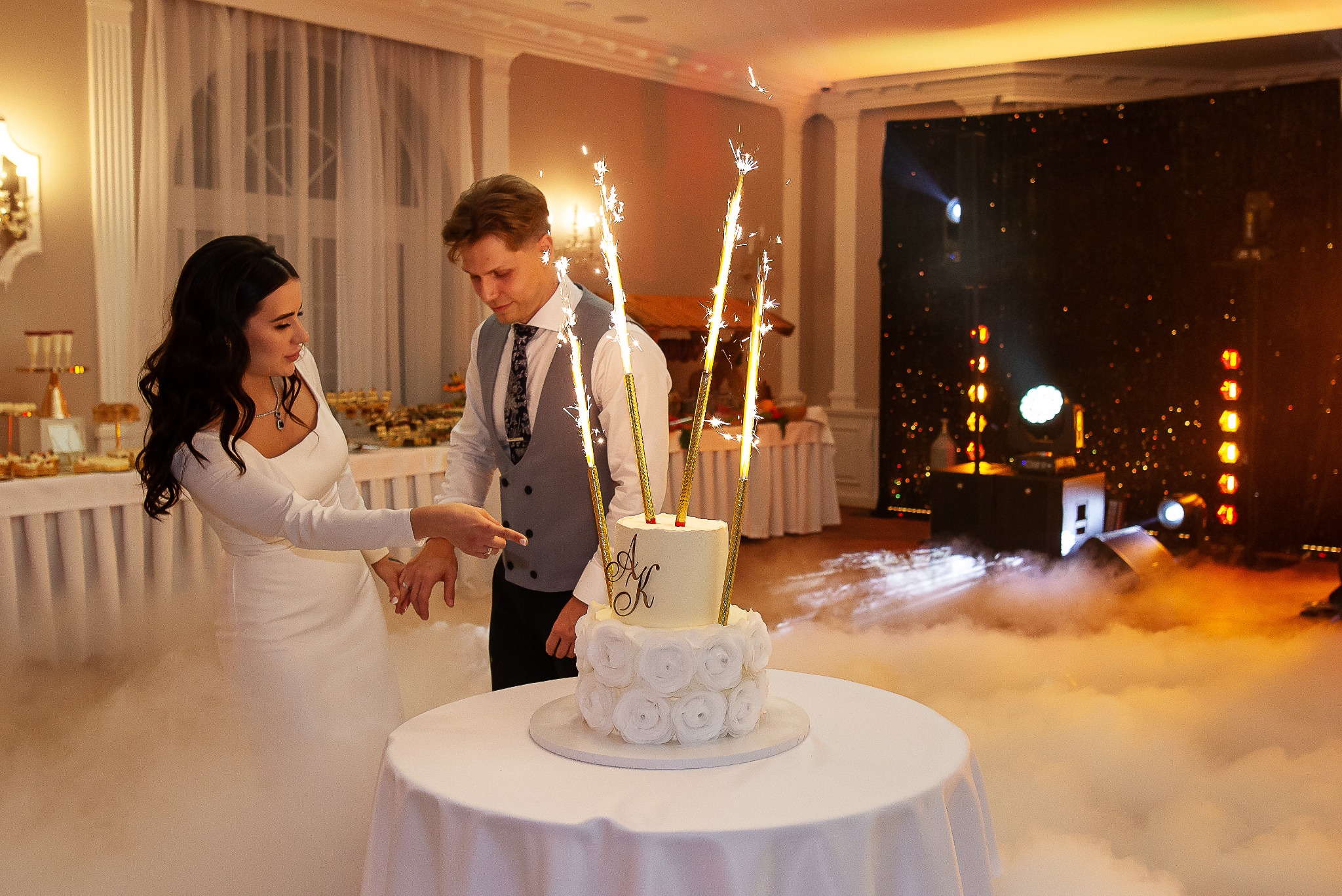 Para młoda kroi tort weselny z fontannami iskier na sali balowej z dekoracjami świetlnymi i efektem dymu. Kobieta w białej sukni wskazuje na tort.