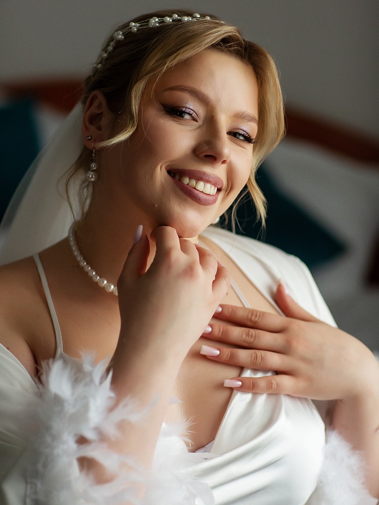 Uśmiechnięta panna młoda w białym szlafroku z piórami, perłowym naszyjnikiem i diademem, z delikatnym makijażem, pozuje do portretu w jasnym wnętrzu.