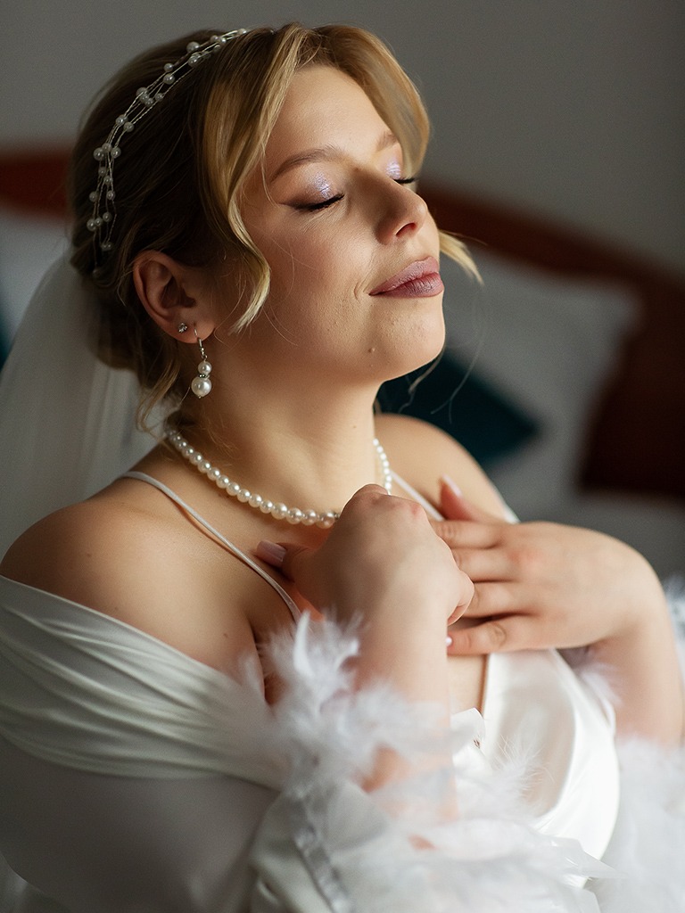 Portret panny młodej w satynowym szlafroku z piórami, z perłami i tiarą, w delikatnym makijażu ślubnym, uchwycony w miękkim świetle w Poznaniu. Zamknięte oczy, spokój.