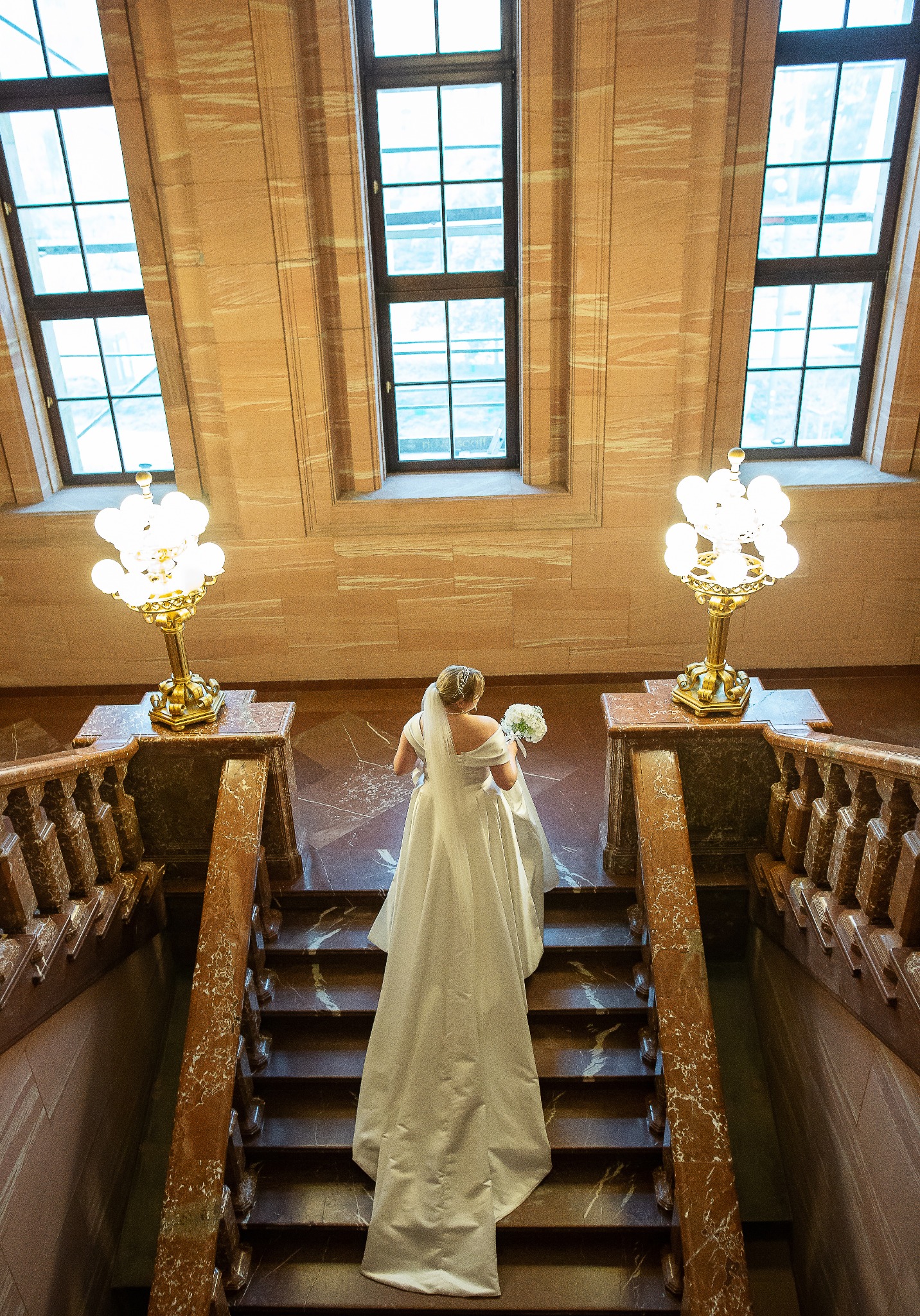 Panna młoda w białej sukni wchodzi po marmurowych schodach, trzymając bukiet. W tle widoczne okna i ozdobne lampy. Ujęcie z góry.