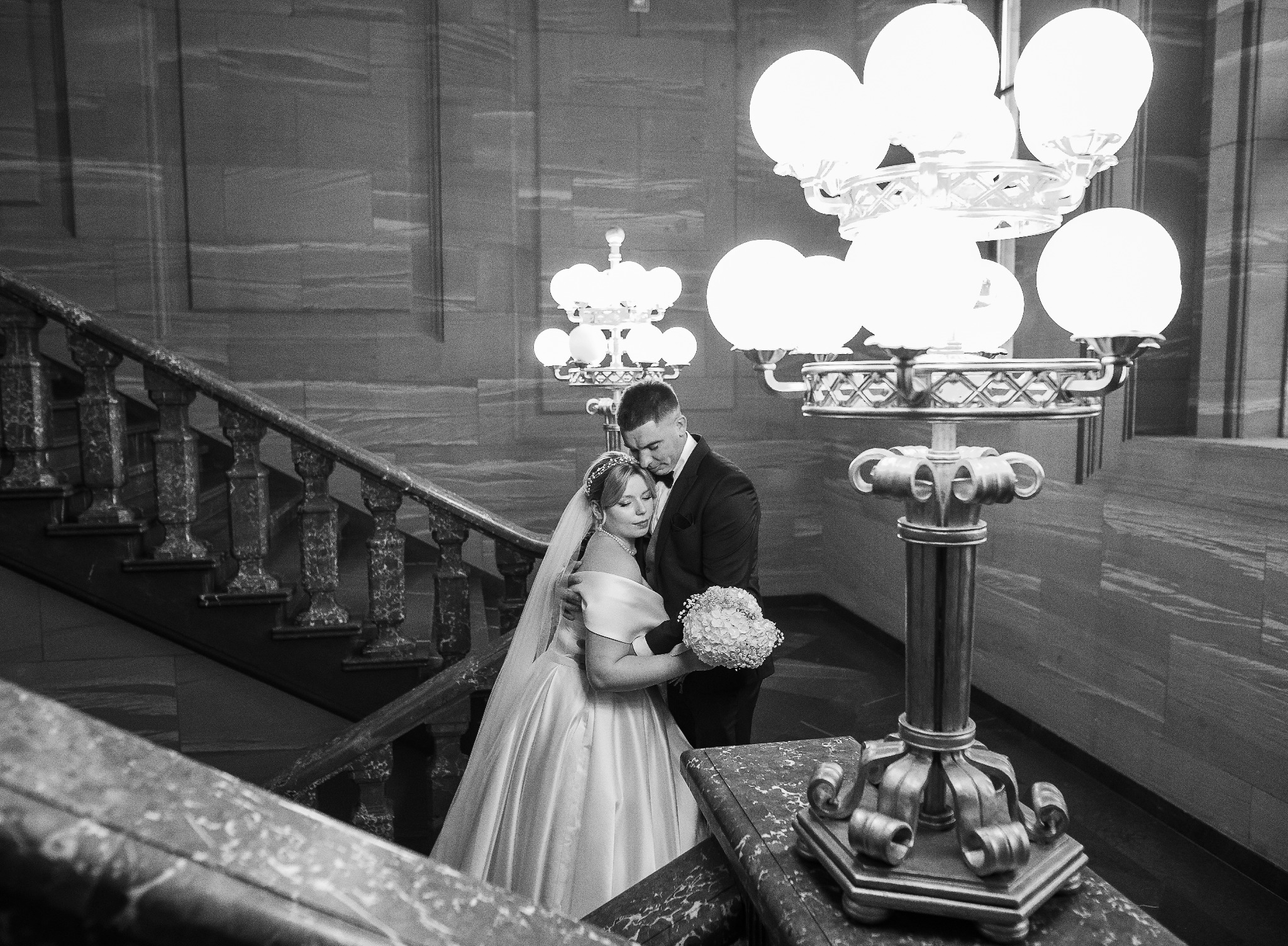 Czarno-białe zdjęcie pary młodej na marmurowych schodach, oświetlonych ozdobnymi lampami. Panna młoda w sukni z bukietem, pan młody w garniturze, obejmujący ją czule.