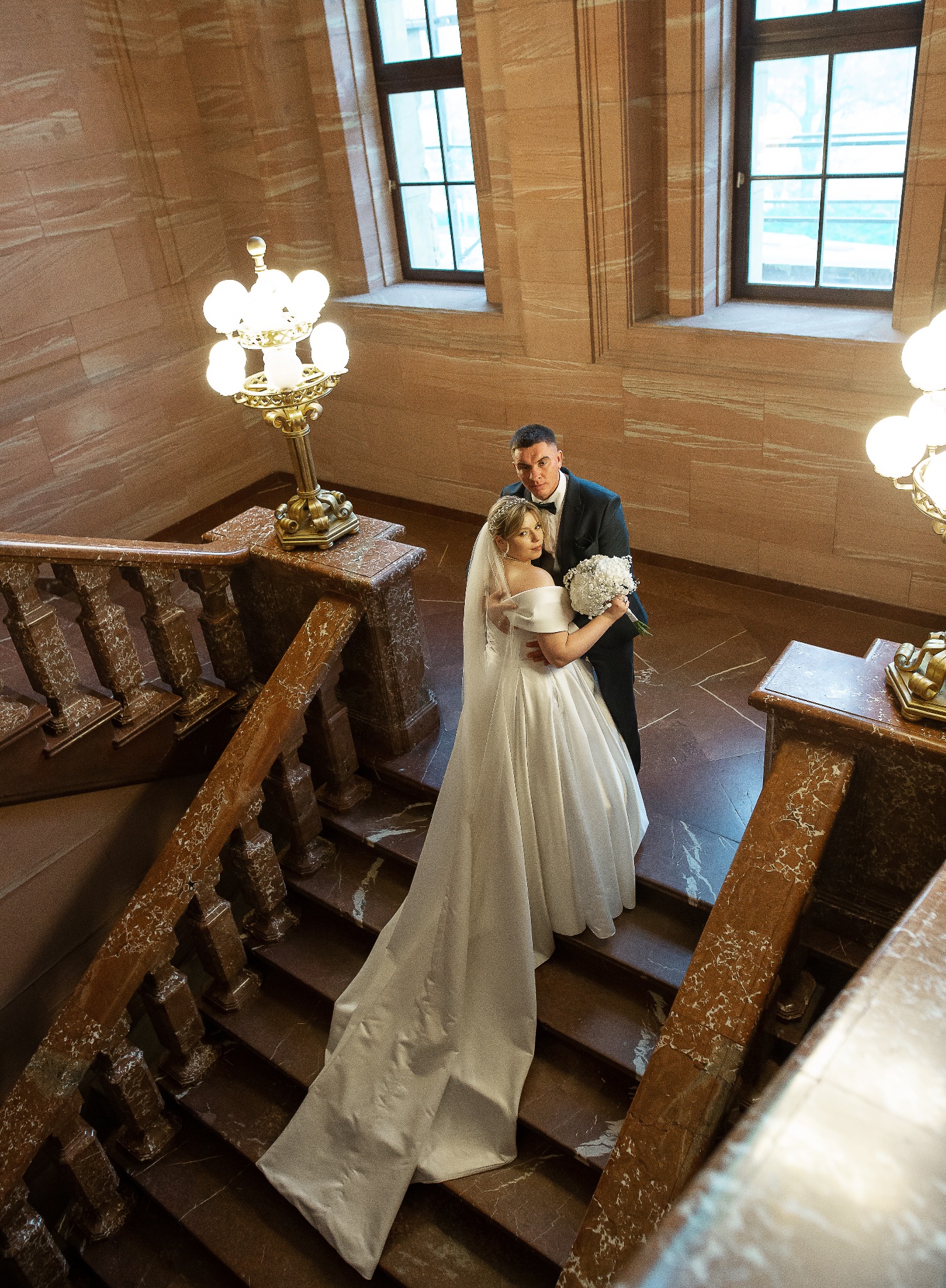 Elegancka para młoda na marmurowych schodach z balustradą, oświetlonych ozdobnymi lampami. Panna młoda w białej sukni z długim trenem, pan młody w czarnym smokingu.