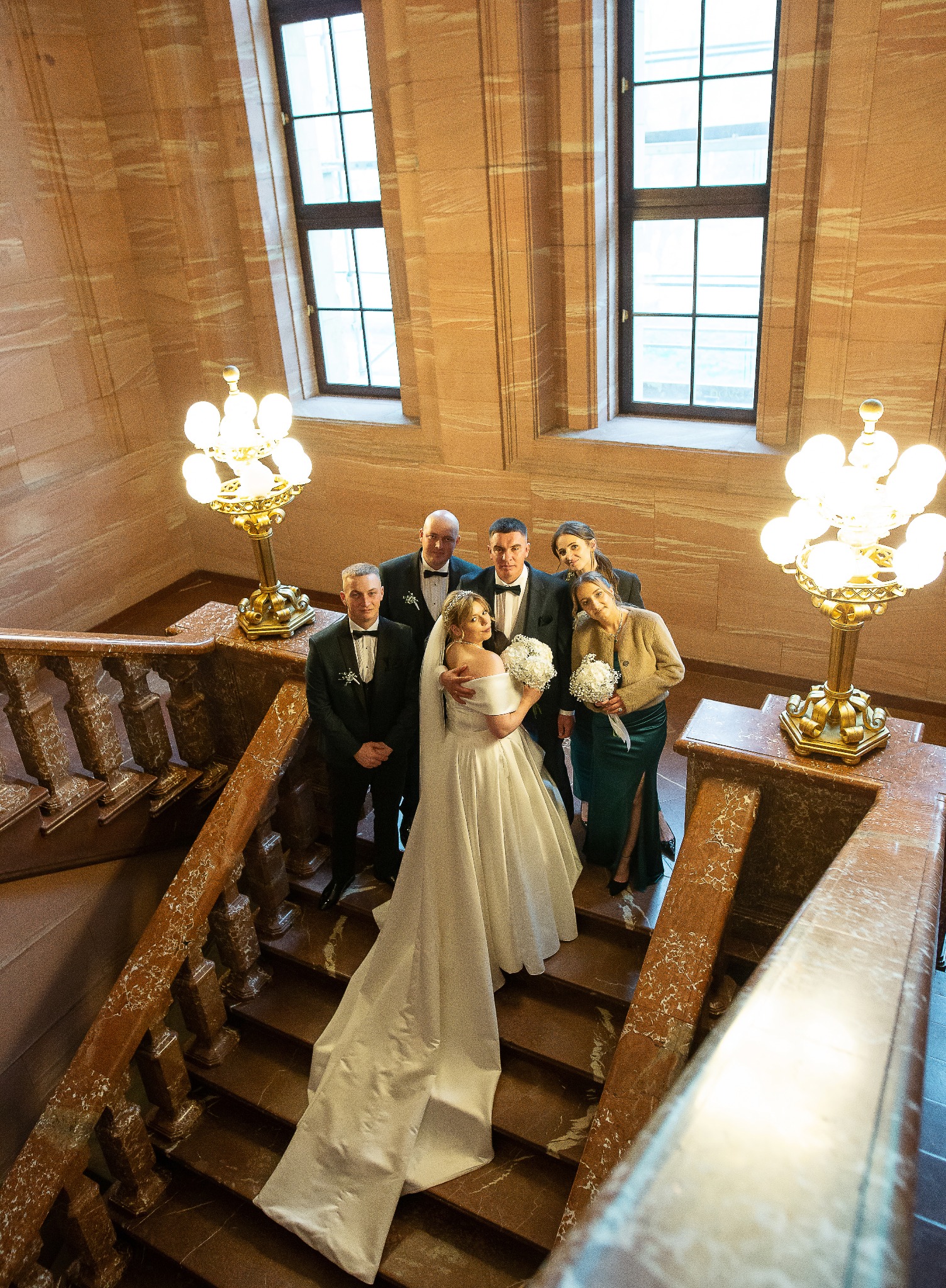Panna młoda w białej sukni i panowie w smokingach na marmurowych schodach z kwiatami, widok z góry, oświetlenie lampami w stylu retro, duże okna w tle.