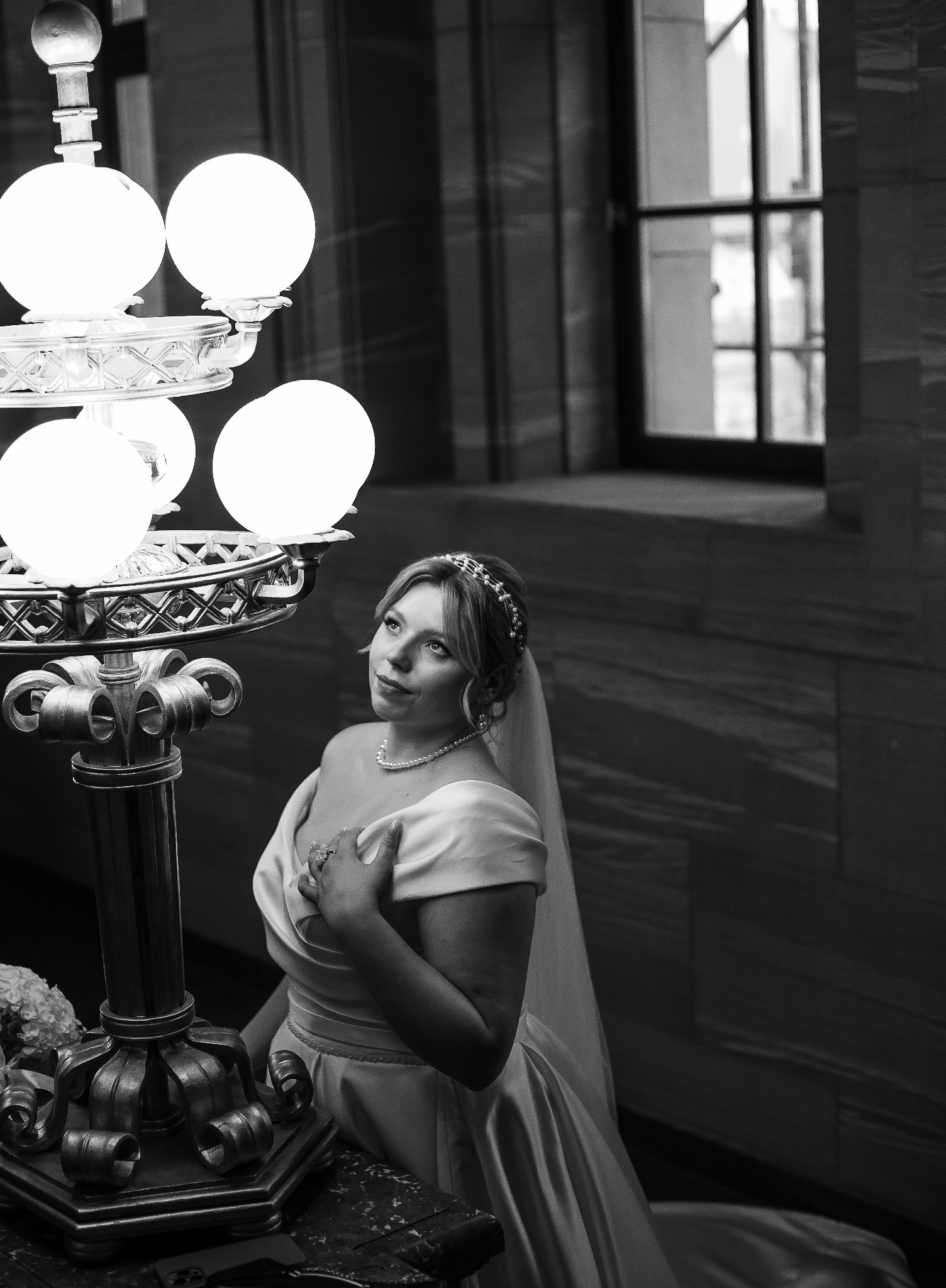 Czarno-białe zdjęcie panny młodej w sukni, z tiarą i perłami, stojącej obok ozdobnej lampy w eleganckim wnętrzu. Spogląda w górę, trzymając dłoń na piersi.