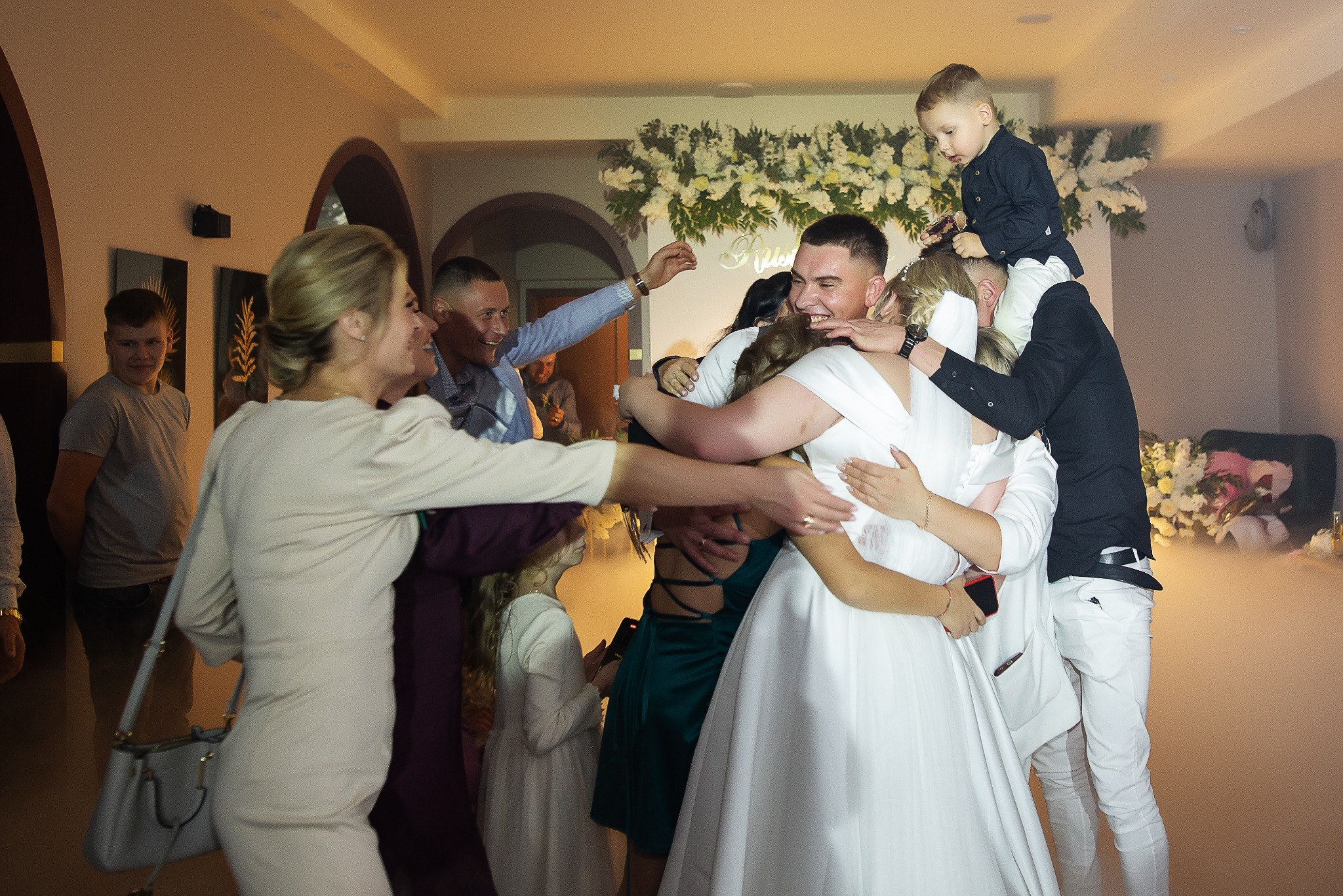 Radosne ujęcie z wesela: para młoda w objęciach gości, chłopiec na barana, dekoracja kwiatowa w tle, ciepłe oświetlenie tworzy uroczystą atmosferę.