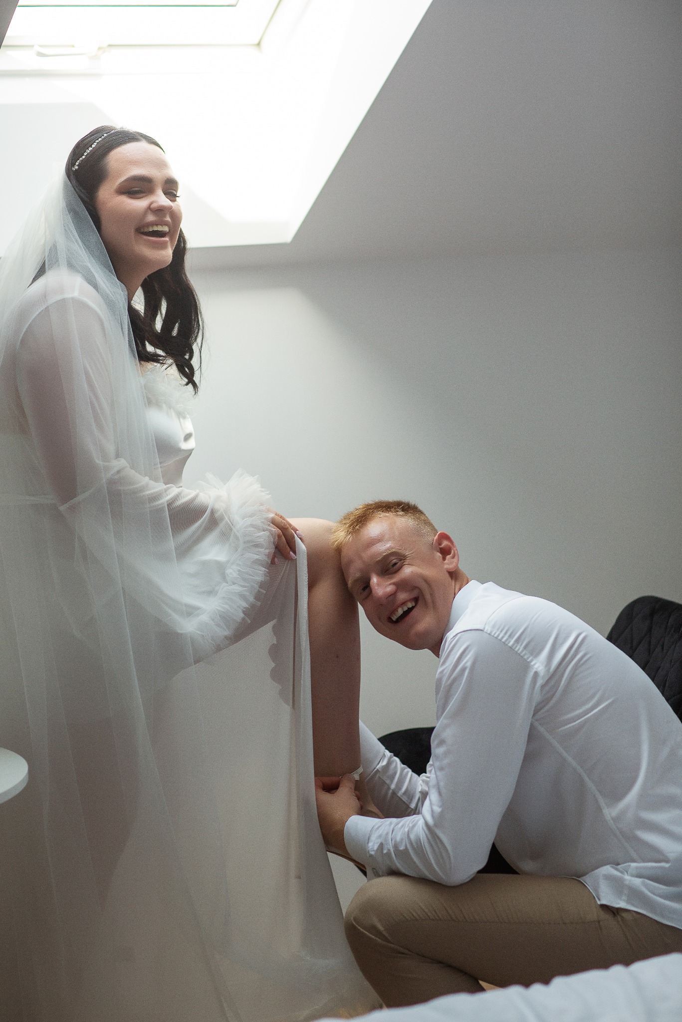 Uśmiechnięta panna młoda w białej sukni i welonie siedzi, opierając nogę na klęczącym mężczyźnie w białej koszuli, oboje patrzą w górę i śmieją się w jasnym pomieszczeniu.