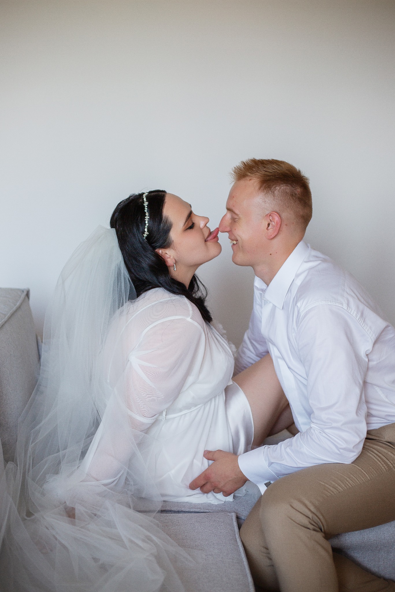 Uśmiechnięta para w jasnych strojach siedzi blisko, dotykając się nosami. Kobieta w welonie i sukience, mężczyzna w koszuli. Delikatne, naturalne światło.