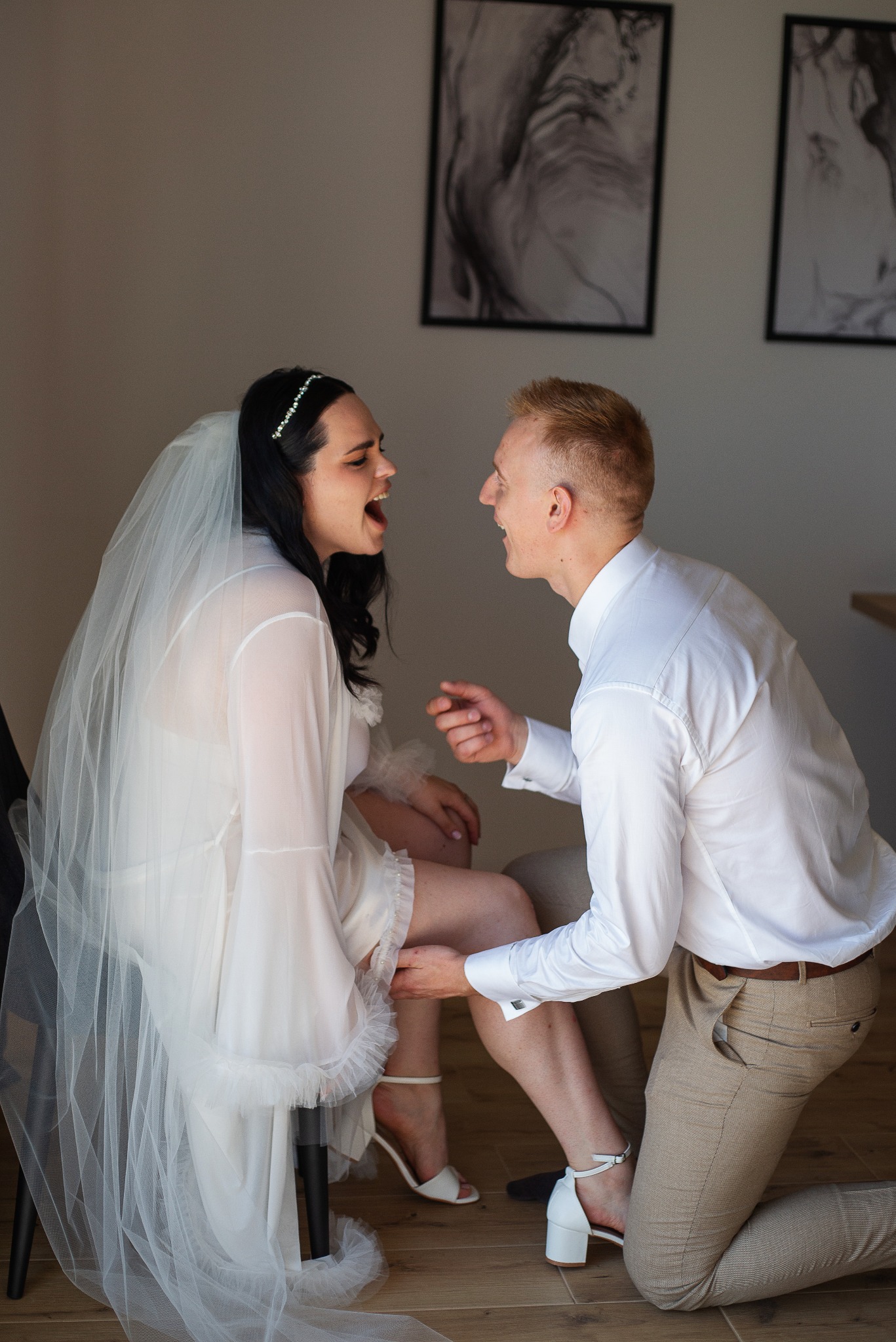 Radosna panna młoda w bieli siedzi, a pan młody klęczy, zakładając jej podwiązkę. Uśmiechnięci, w jasnym wnętrzu z minimalistycznymi obrazami w tle.