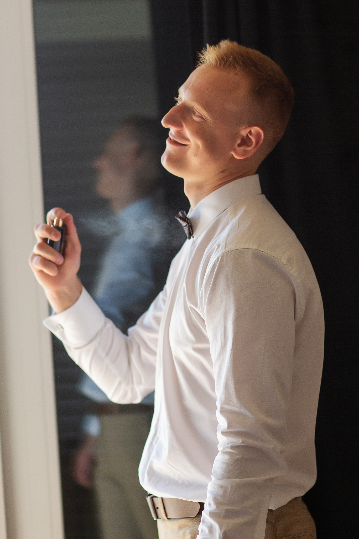 Uśmiechnięty mężczyzna w białej koszuli i muszce, profil, aplikuje perfumy. W tle widoczne odbicie w lustrze i czarna zasłona.