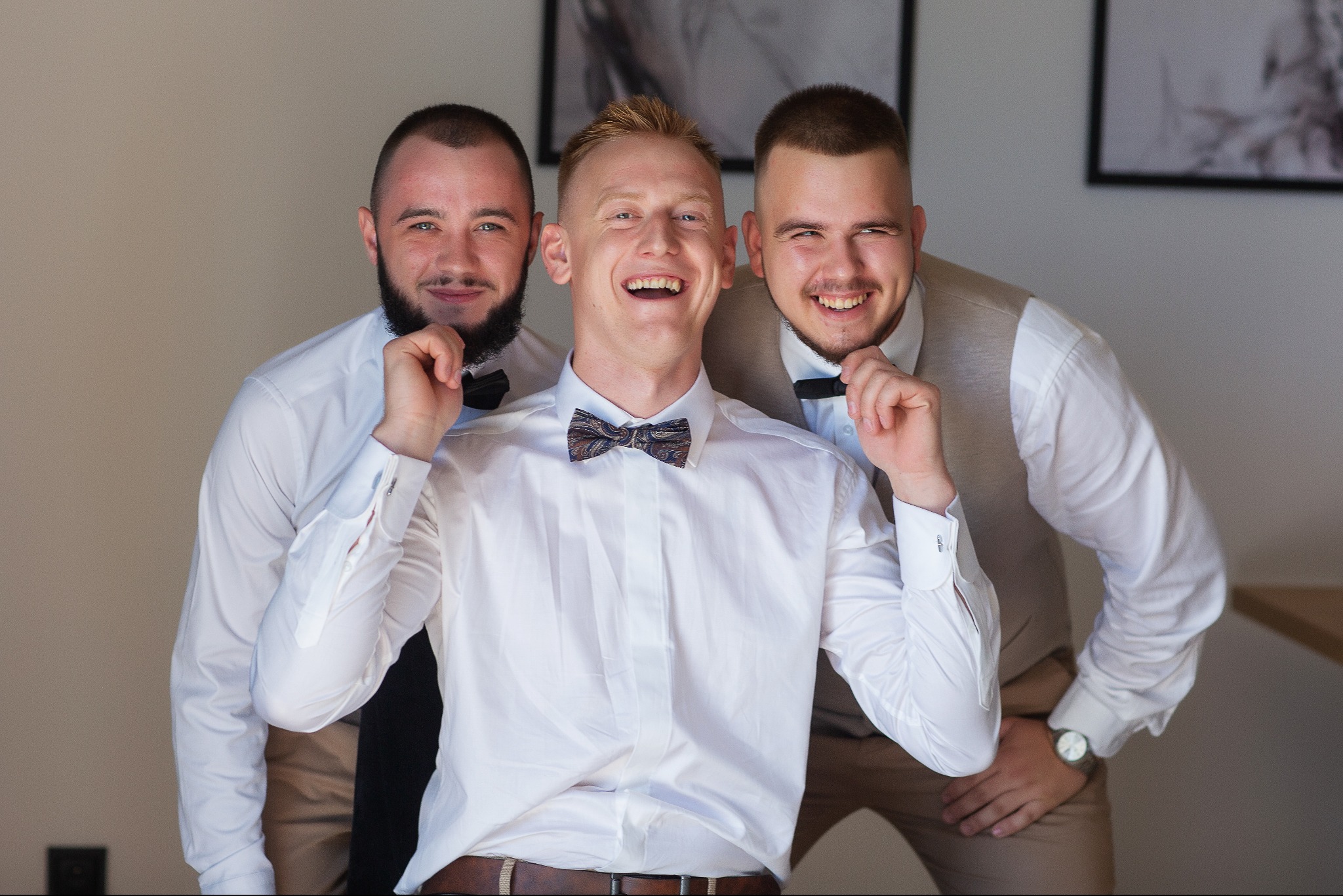 Trzech uśmiechniętych mężczyzn w białych koszulach i muchach, pozujących do zdjęcia w jasnym wnętrzu. Środkowy mężczyzna ma wzorzystą muchę.