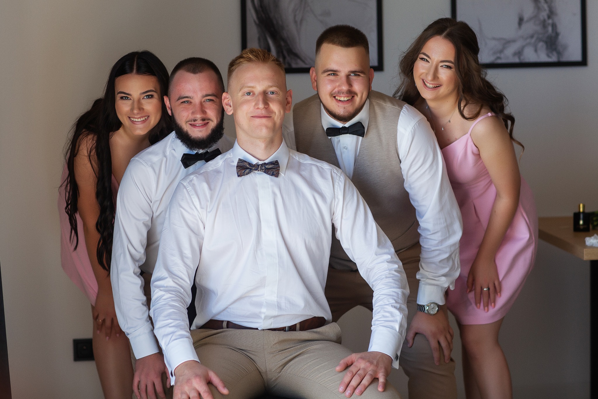 Grupowe zdjęcie uśmiechniętych osób w eleganckich strojach, w jasnym wnętrzu. Wykonane w Poznaniu, idealne na sesję narzeczeńską lub biznesową.