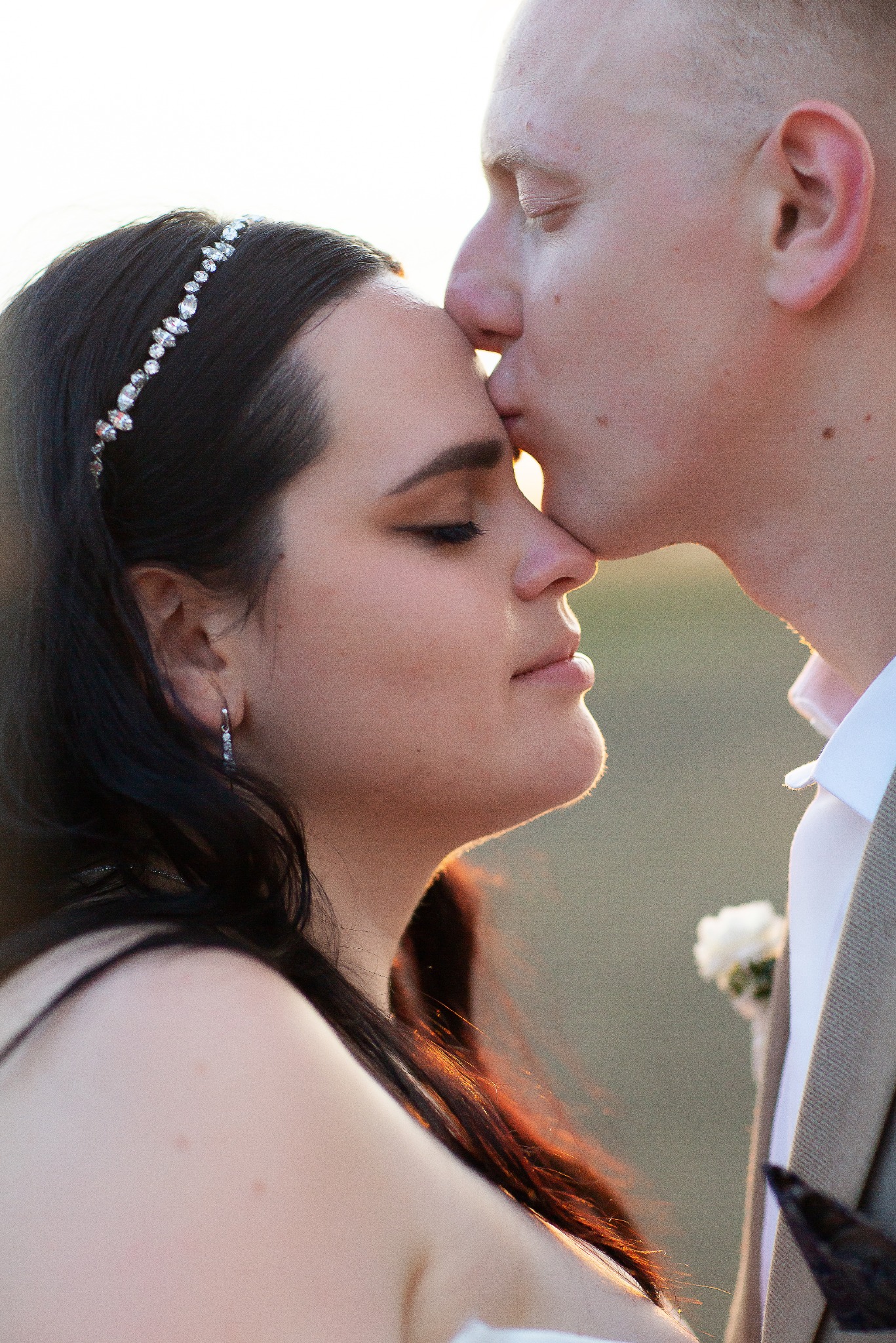 Profil narzeczonej z diademem, całowanej w czoło przez narzeczonego w beżowym garniturze, w ciepłym świetle zachodzącego słońca. Delikatne kolczyki i kwiat w butonierce.