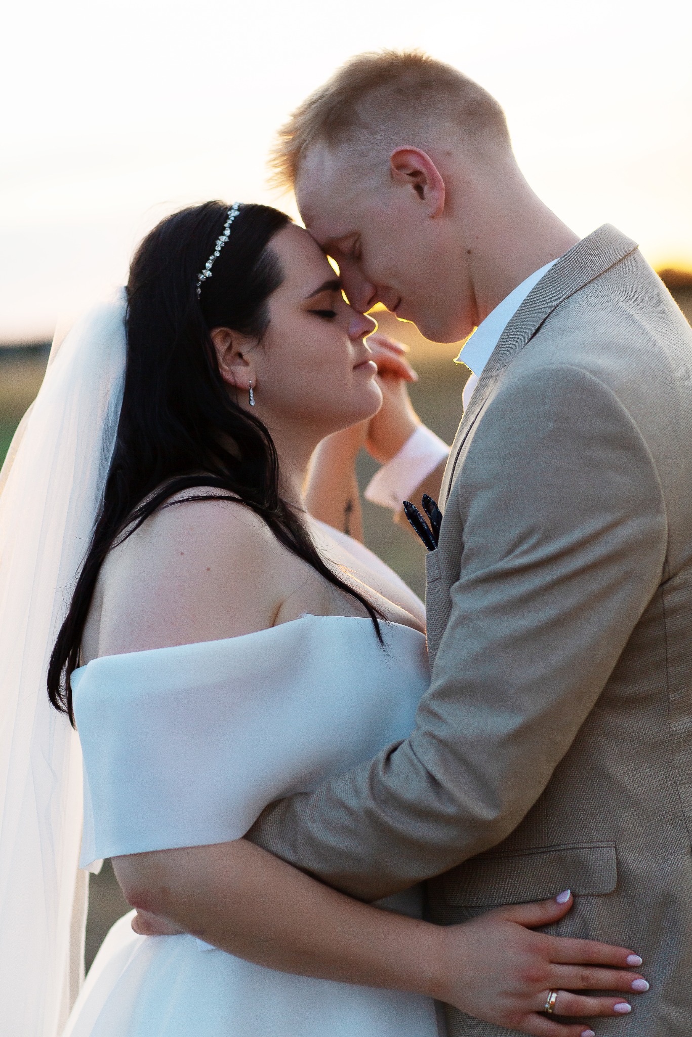 Młoda para w objęciach, panna młoda w białej sukni i welonie, pan młody w beżowym garniturze, zbliżenie twarzy, ciepłe światło zachodzącego słońca w tle.