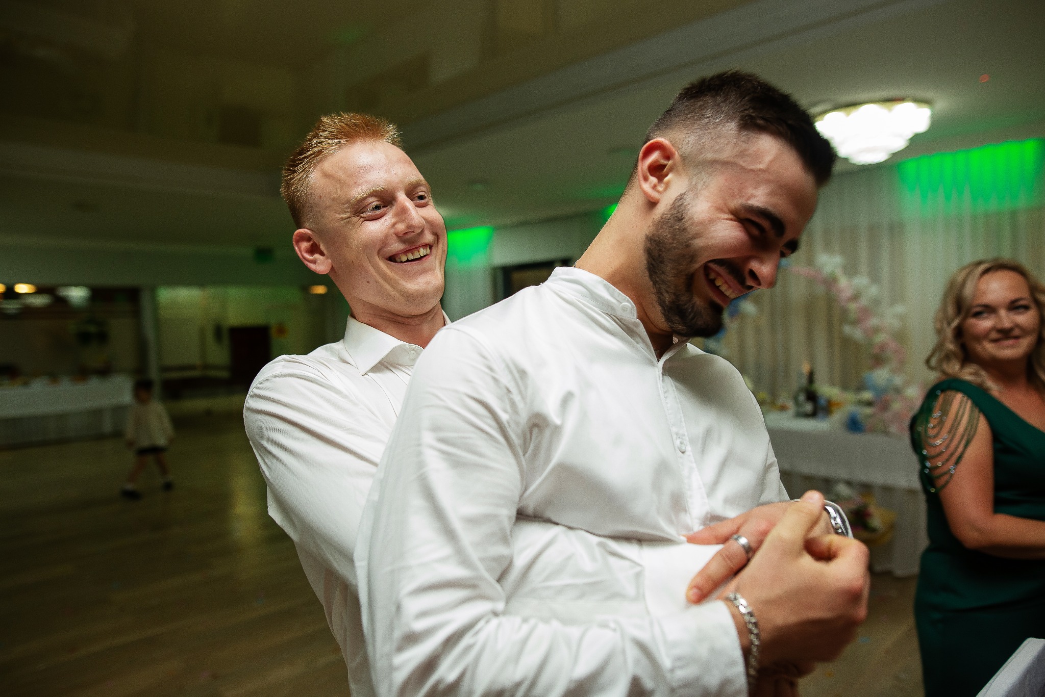Radosne ujęcie dwóch mężczyzn w białych koszulach, śmiejących się podczas uroczystości. W tle widoczny stół z dekoracjami i kobieta w zielonej sukience.