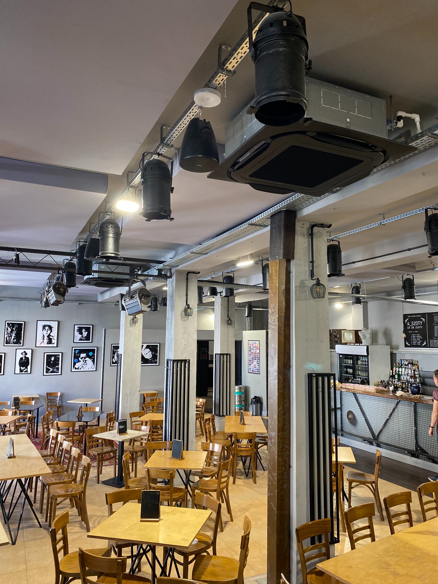 Wnętrze lokalu gastronomicznego z widoczną kasetonową klimatyzacją sufitową, oświetleniem punktowym na szynoprzewodach, stolikami i krzesłami oraz barem w tle.