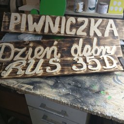 Mirosław Gielka - Dwie drewniane tabliczki z wyciętymi napisami 'Piwniczka' i 'Dzień dobry RUS 35D', z wypalonym tłem, leżące na warsztatowym stole, w tle puszki z farbami i zielona wycinanka w kształcie żaby.
