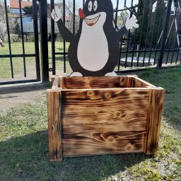 Mirosław Gielka - Opalana drewniana skrzynia na rośliny z dekoracją w postaci postaci Krecika, umieszczona na trawniku przed metalowym ogrodzeniem.