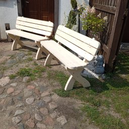 Mirosław Gielka - Dwie surowe, jasne, drewniane ławki ogrodowe z krzyżowymi nogami stoją na brukowanym podwórku przed domem z ciemnymi drzwiami i ozdobnymi roślinami.